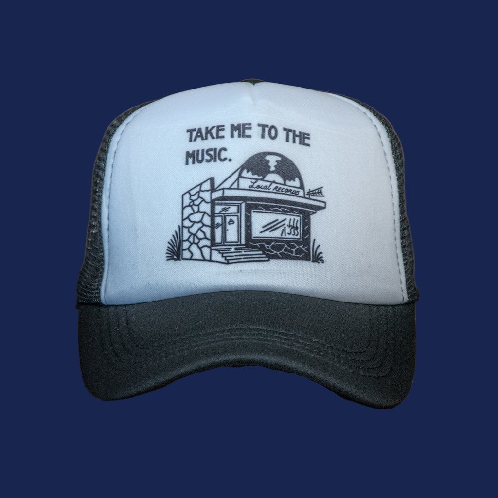 Black and white cap with 'Take Me to the Music' text and graphic on a wooden surface.